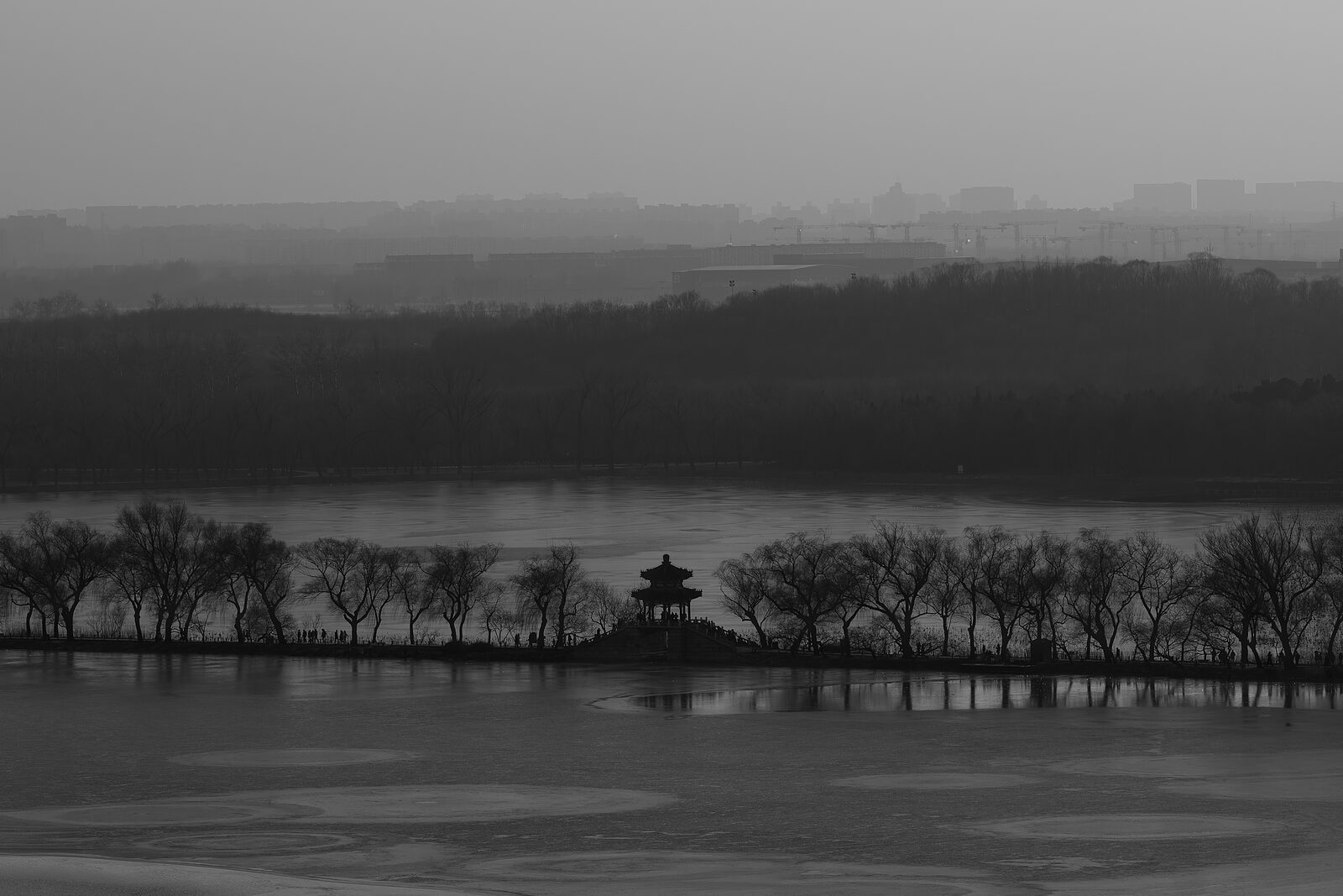 A small pavilion on the far bank of a frozen lake in monochrome, the Beijing skyline dissolving into winter haze — everything still gray
