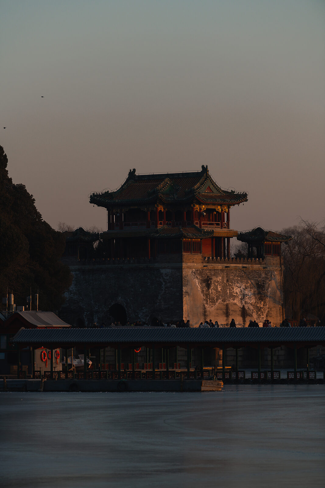 A gate tower above the lake at blue dusk, the last warm light on its weathered wall, people gathered along the covered walkway