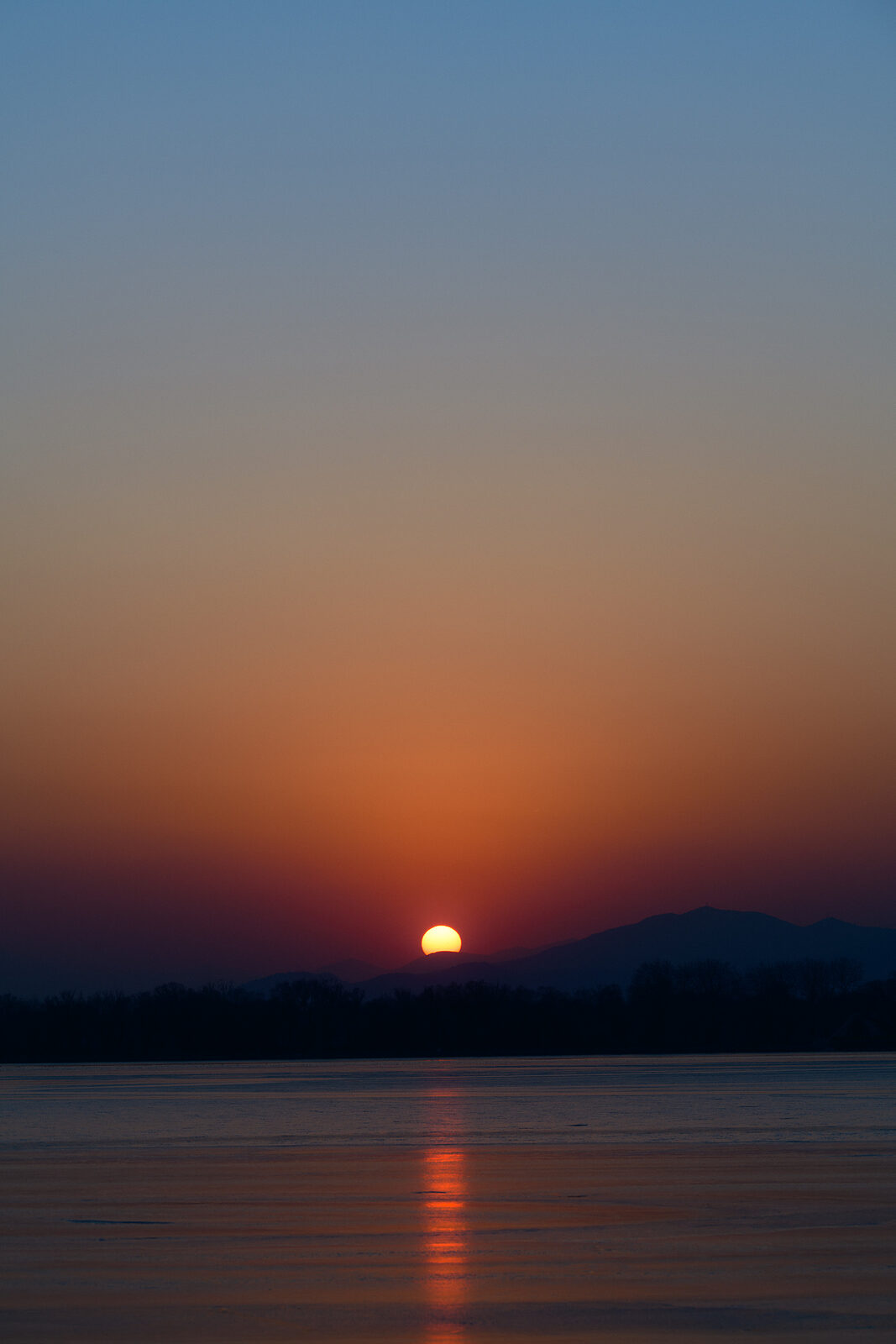 The winter sun sinking behind distant mountains, its light trailing in a thin line across the frozen lake