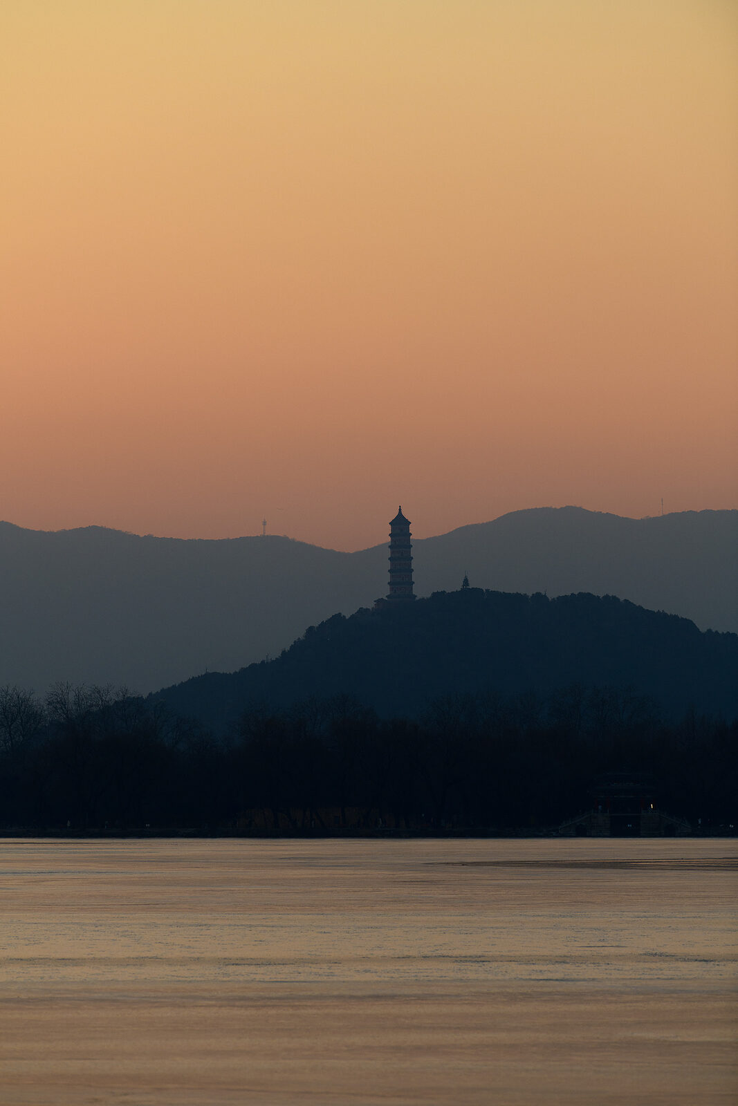 A slender pagoda on a distant wooded hill, silhouetted against an amber dusk sky above the frozen lake