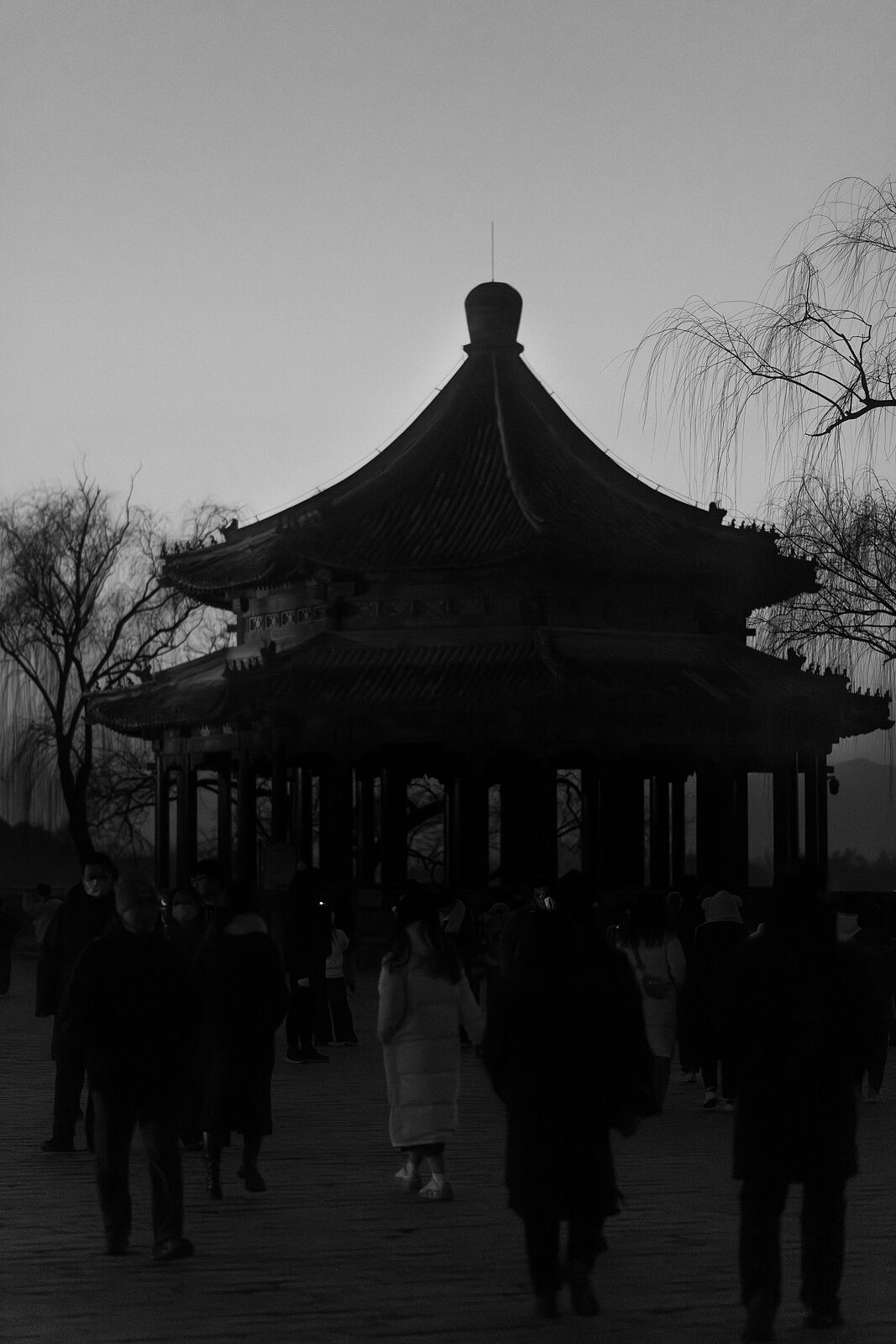 Visitors moving as dark silhouettes around a pavilion at nightfall, rendered in black and white