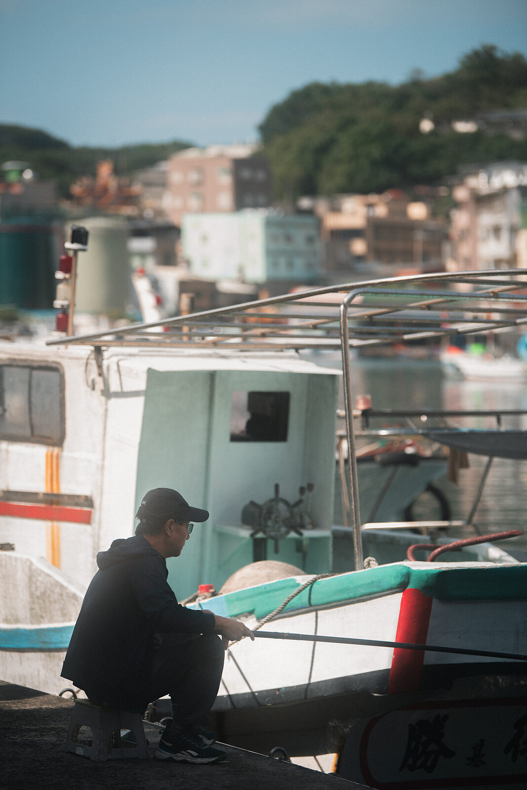 A man in black crouching at the harbor edge, fishing line held between his hands, boats resting behind him