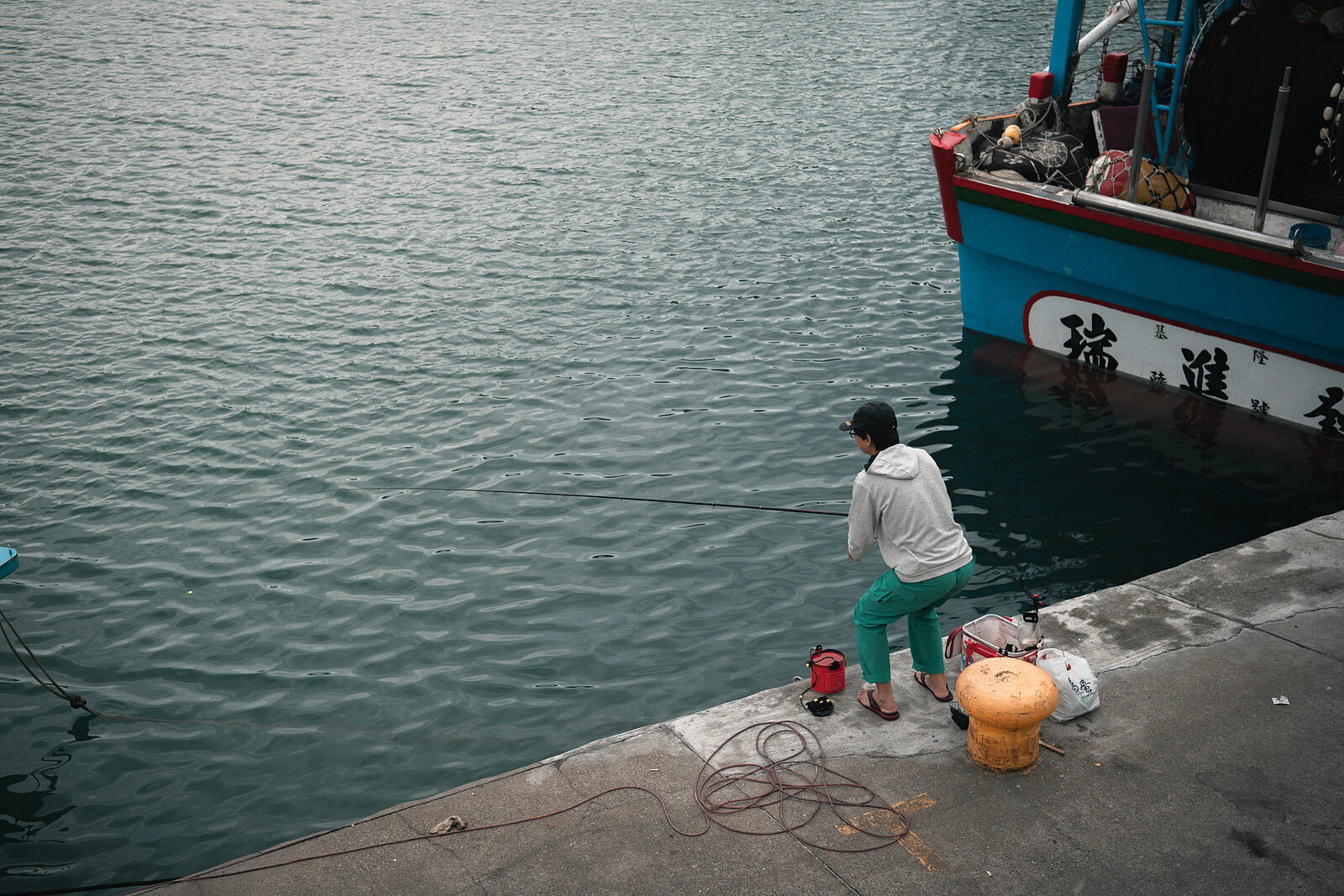 A figure in white seated on the pier, rod extended over grey water, a Taiwanese fishing boat painted blue beside her