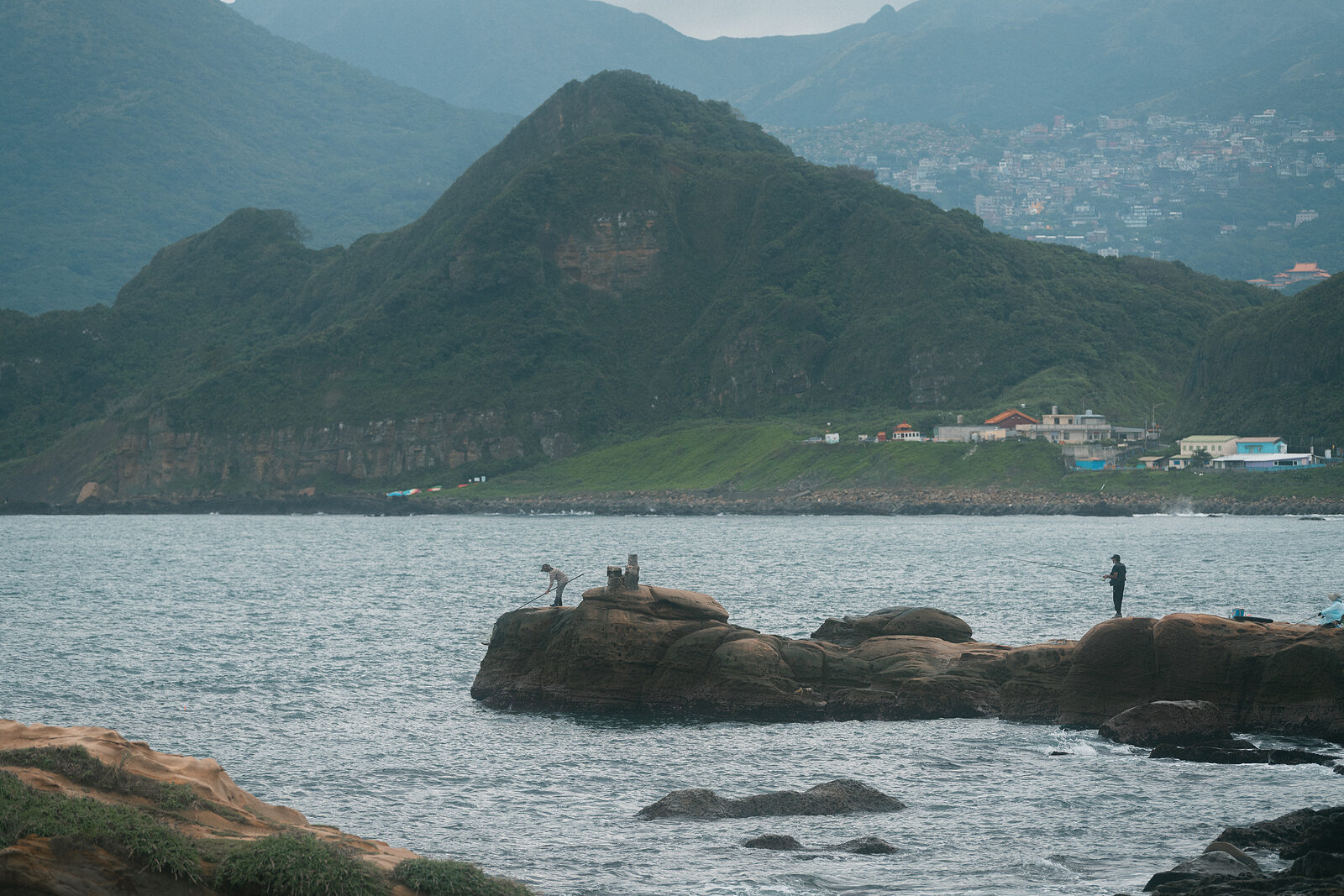 Two small figures on coastal rocks, casting rods into the sea, green mountains disappearing into mist behind them