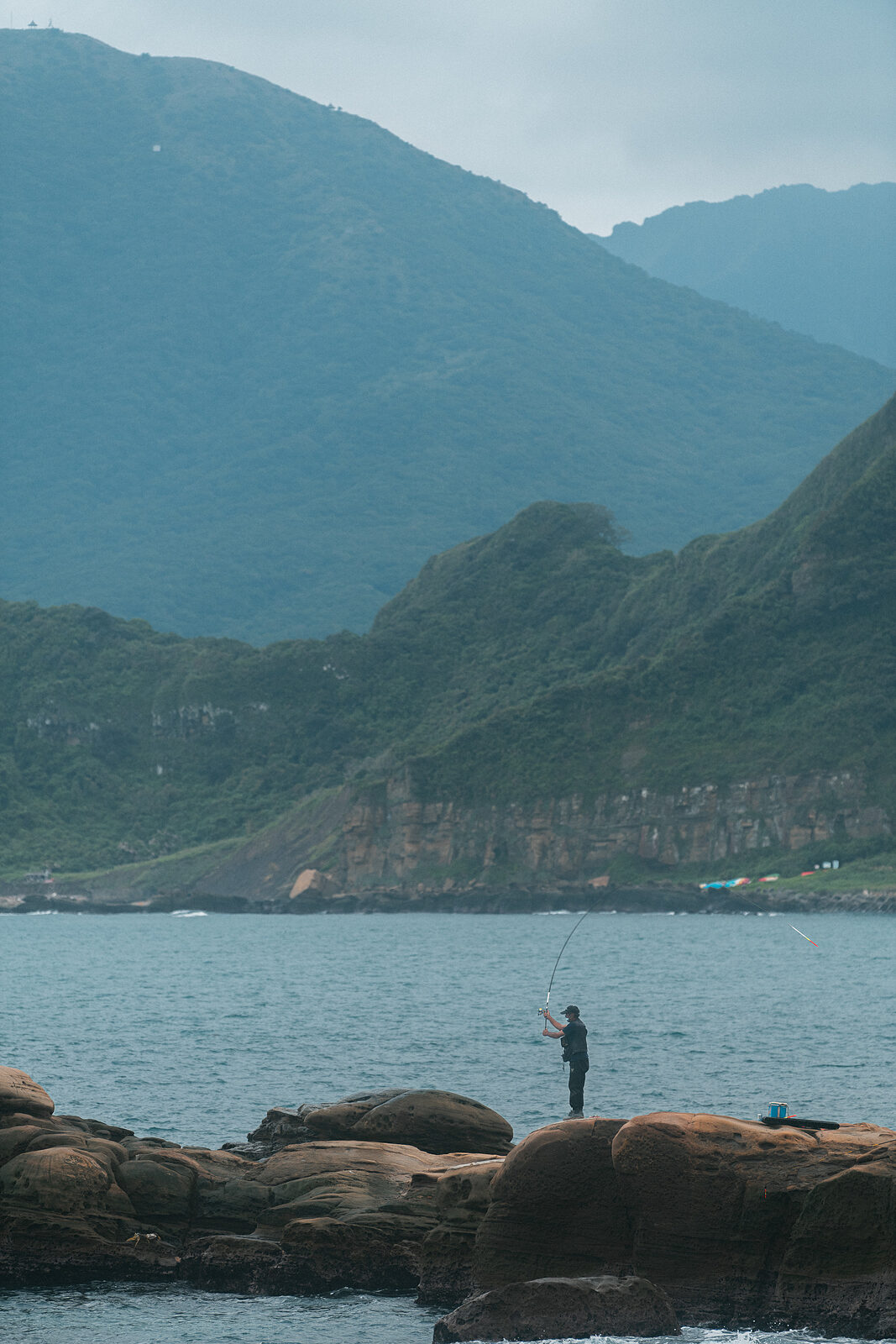 A lone fisherman standing on a boulder, rod raised in an arc against the mountain and overcast sky