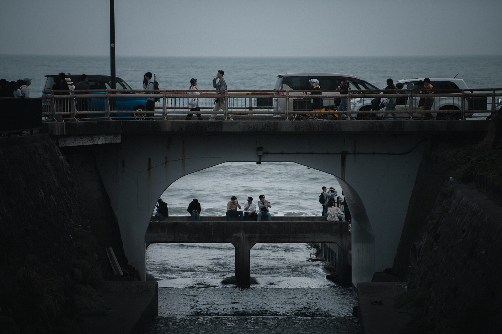 A bridge by the sea, people passing across the top and others seated beneath the arch