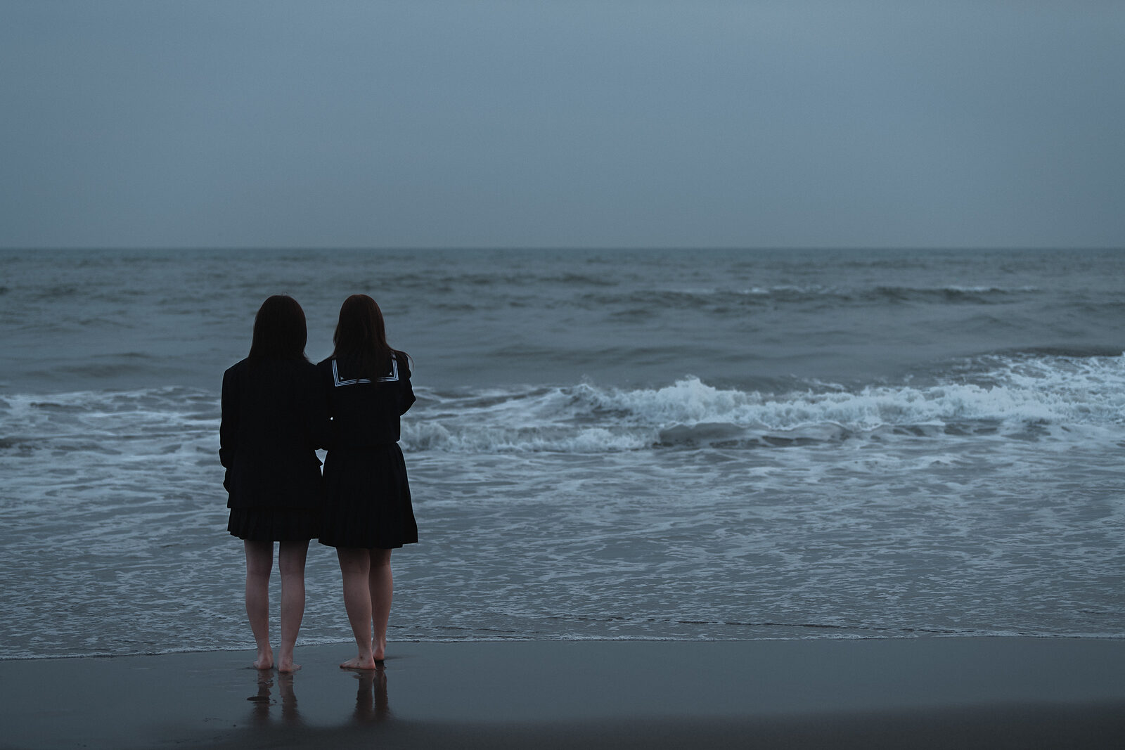 Two students in school uniforms standing barefoot at the edge of the ocean