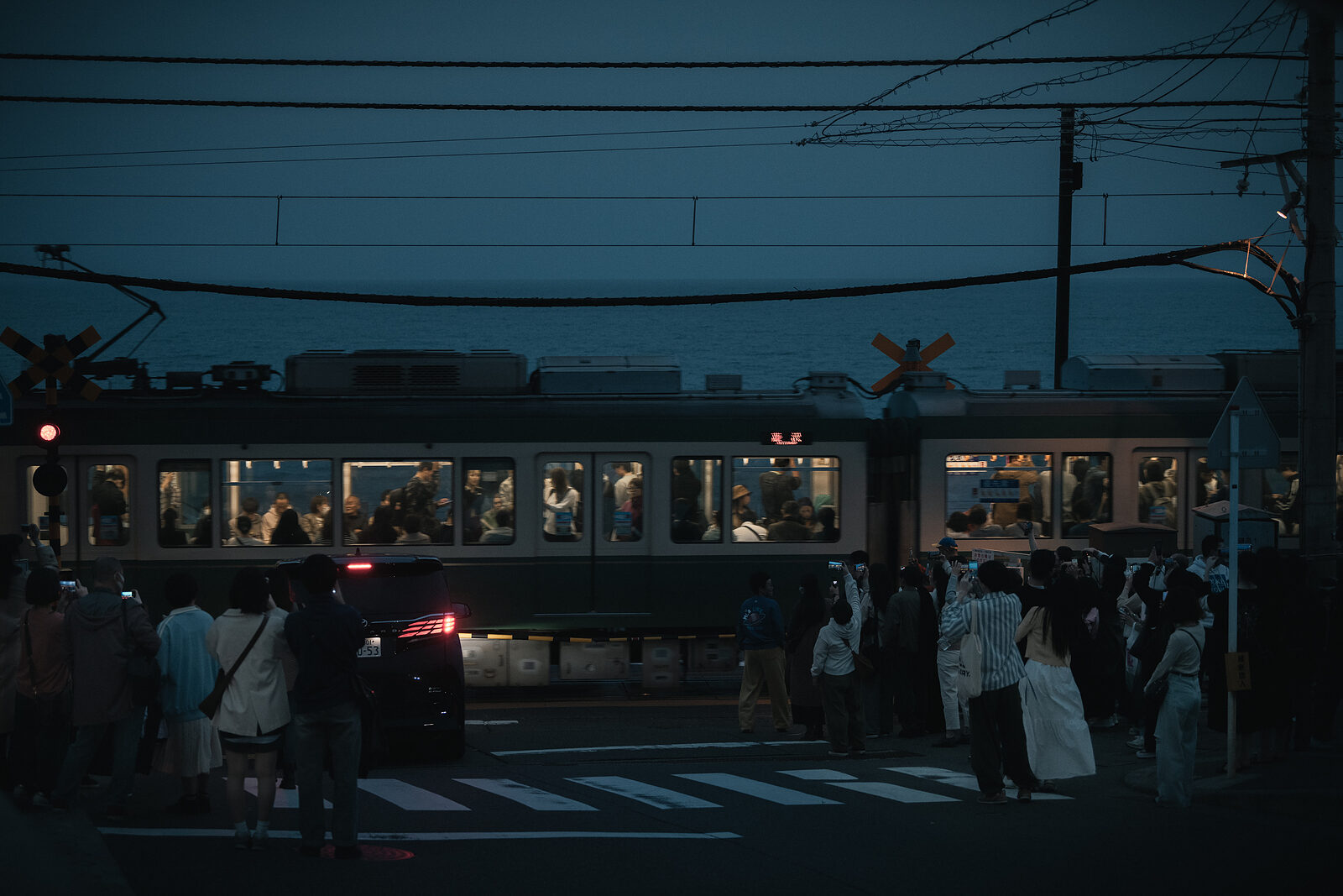 An Enoden train passing through a crossing at dusk, surrounded by spectators with phones raised